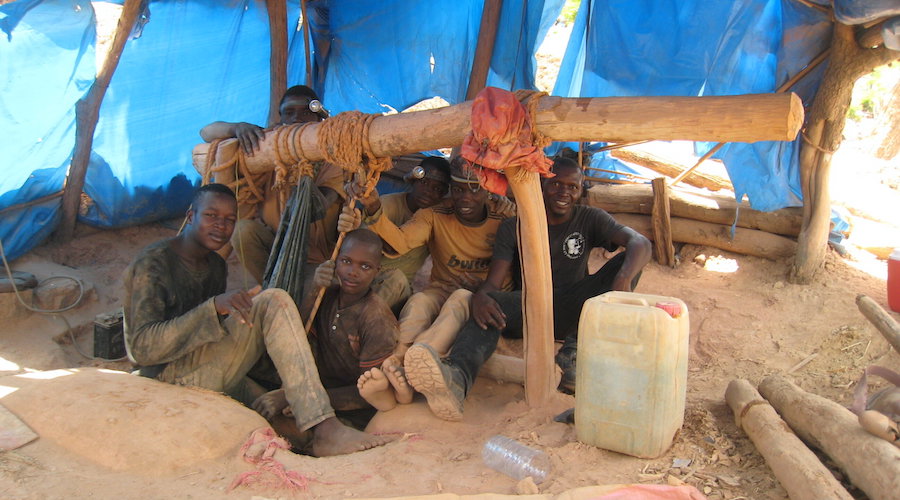 Miners pose for a photo near a dug-out mining pit under a blue tarp held up by a frame made from logs and branches.